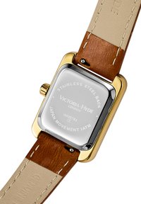 Montre en acier inoxydable doré avec un boîtier rectangulaire, bracelet en cuir marron et dessous beige clair. Présente des détails de marque et de mouvement gravés.