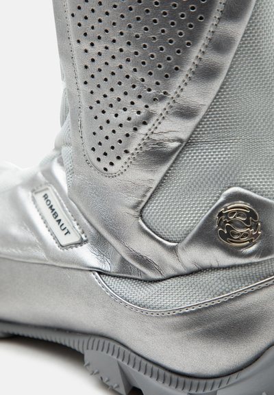 Κοντινή λήψη ασημένιας μπότες Rombaut που δείχνει διάτρητο δέρμα, υφασμάτινη επιφάνεια με υφή, μεταλλικό λογότυπο και παχύ πέλμα από καουτσούκ.