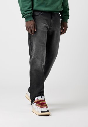 Person trägt ausgebleichte schwarze Jeans und weiße Sneakers mit dicken, mehrfarbigen gelb-roten Schnürsenkeln, steht vor einfachem Hintergrund.