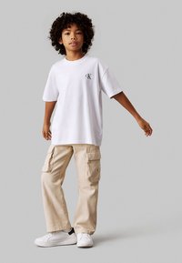 Balta kokvilnas T-krekls ar mazu pelēku logotipu, kopā ar bēšām biksēm ar sānu kabatām un logotipa akcentiem. Valkāts ar baltām sporta kurpēm.