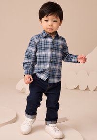 Kék árnyalatú, fehér részletekkel díszített kockás ing, sötét farmernadrág és fehér tornacipő. Az ingnél medve hímzés látható.