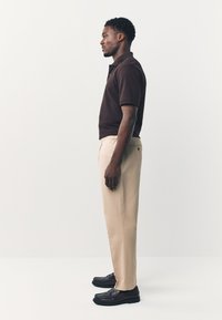 Braunes Poloshirt, beige Hose und schwarze Schuhe. Das Modell steht im Profil und präsentiert eine lockere Passform mit kurzen Ärmeln und klassischem Kragen.