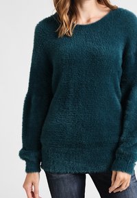 Teal fluffig tröja med rund halsringning, långa ärmar och ribbade muddar. Mjuk textur och avslappnad passform; synlig fåll vid höfterna.