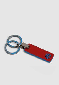 Rode sleutelhangerlabel met blauwe accenten, bevestigd aan twee zilveren metalen ringen. Heeft een gladde textuur en een verhoogd logo op het label.