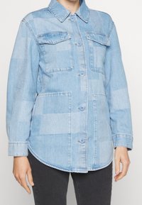 Světle modrá džínová košile s jaklovým vzorem, dvěma náprsními kapsami, zakulaceným lemem a knoflíkovým zapínáním. Hladká textura, ležérní design.