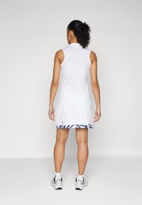 White sleeveless athletic dress with a high collar, featuring a navy patterned trim at the hem and side slits. Worn with gray sports shoes.