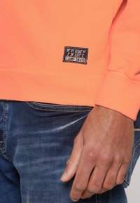 Sudadera naranja con ribete en el bajo y los puños, que presenta una etiqueta negra con coordenadas y el nombre de la marca "CAMP DAVID." Llevada con jeans de mezclilla azul.