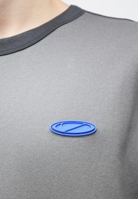 Γκρι βαμβακερό T-shirt με μπλε ελαστικό οβάλ έμβλημα στο αριστερό μέρος του στήθους με ανάγλυφο σχέδιο. Ριμ�π λαιμός. Μαλακή υφή.