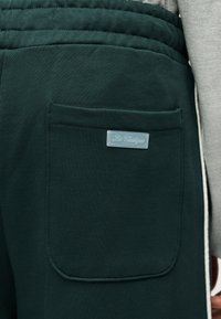 Κοντινή λήψη τσέπης σκούρου πράσινου παντελονιού φόρμας με μικρή ετικέτα «Les Classiques», που φοριέται μαζί με ένα ανοιχτό γκρι μανίκι ορατό.