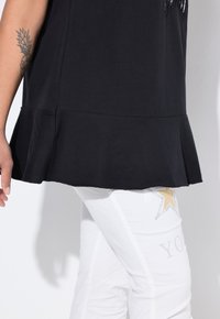 Černý bavlněný top s rozšířeným lemem, v kombinaci s bílými kalhotami se zlatou hvězdou a kamínkovými detaily. Detailní pohled na outfit.