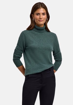 Un pull à col roulé vert foncé en tissu tricoté texturé, avec des manches longues et des poignets côtelés, associé à un pantalon noir.