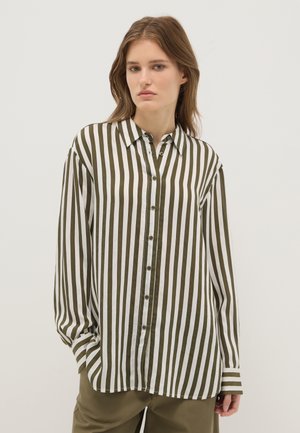 Jeune femme portant une chemise à boutons ample avec des rayures verticales olive et blanches, et un pantalon olive, sur un fond clair uni.