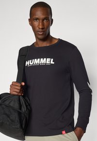 Juodas ilgomis rankovėmis sportinis marškinėliai su „HUMMEL“ logotipu balta spalva, apvaliu kaklu ir pilkiais rankovių akcentais, derinami su juodu sportiniu krepšiu.