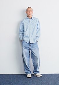 Blauw-wit gestreepte blouse met lange mouwen, lichte spijkerbroek, witte sneakers, staand tegen een effen muur met handen in de zakken.