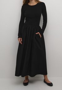 Czarna sukienka z długim rękawem, z prążkowanym korpusem i zwiewnym dołem. Posiada marszczoną talię oraz boczne kieszenie. Dopasowana z czarnymi balerinami.