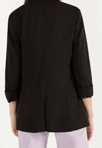 Svart formell kavaj med uppvikt ärmar, skräddarsydd passform och ventilerad baksida. Slätt tyg och inga synliga metalldetaljer.