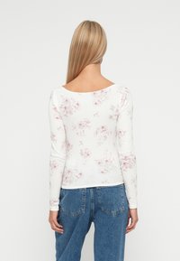 Langarmshirt aus weißer Baumwolle mit einem Blumenmuster in Rosa und Beige. Verfügt über einen weiten Ausschnitt und eine gerippte Textur. Getragen mit blauen High-Waist-Jeans.