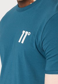 Person som bär en turkosblå crewneck T-shirt med en vit "11°"-logotyp på bröstet, som visar en del av halsen, axeln och armen.