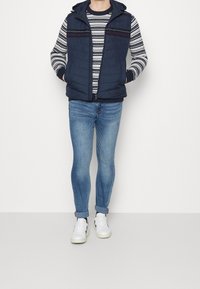 Marineblaue gepolsterte Weste mit Kapuze über einem gemusterten Pullover und schmal geschnittenen blauen Jeans, vervollständigt mit weißen Turnschuhen. Strukturiertes Material.