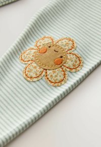 Tissu à rayures bleu clair orné d'un grand patch de fleur texturé avec un visage souriant, agrémenté de coutures orange et de détails floraux.