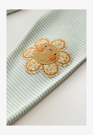 Material în dungi albastru deschis prezintă o aplicație mare și texturată în formă de floare cu o față zâmbitoare, împodobită cu cusături portocalii și detalii florale.