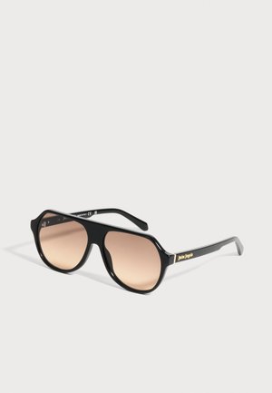 Lunettes de soleil aviateur à monture noire, verres teintés marron et marquage doré "Palm Angels" sur les branches, exposées sur une surface blanche.