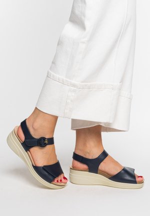 Füße, die marineblaue, offene Keilsandaletten mit Knöchelriemen und Schnallen tragen, kombiniert mit weißen, gekürzten Hosen.