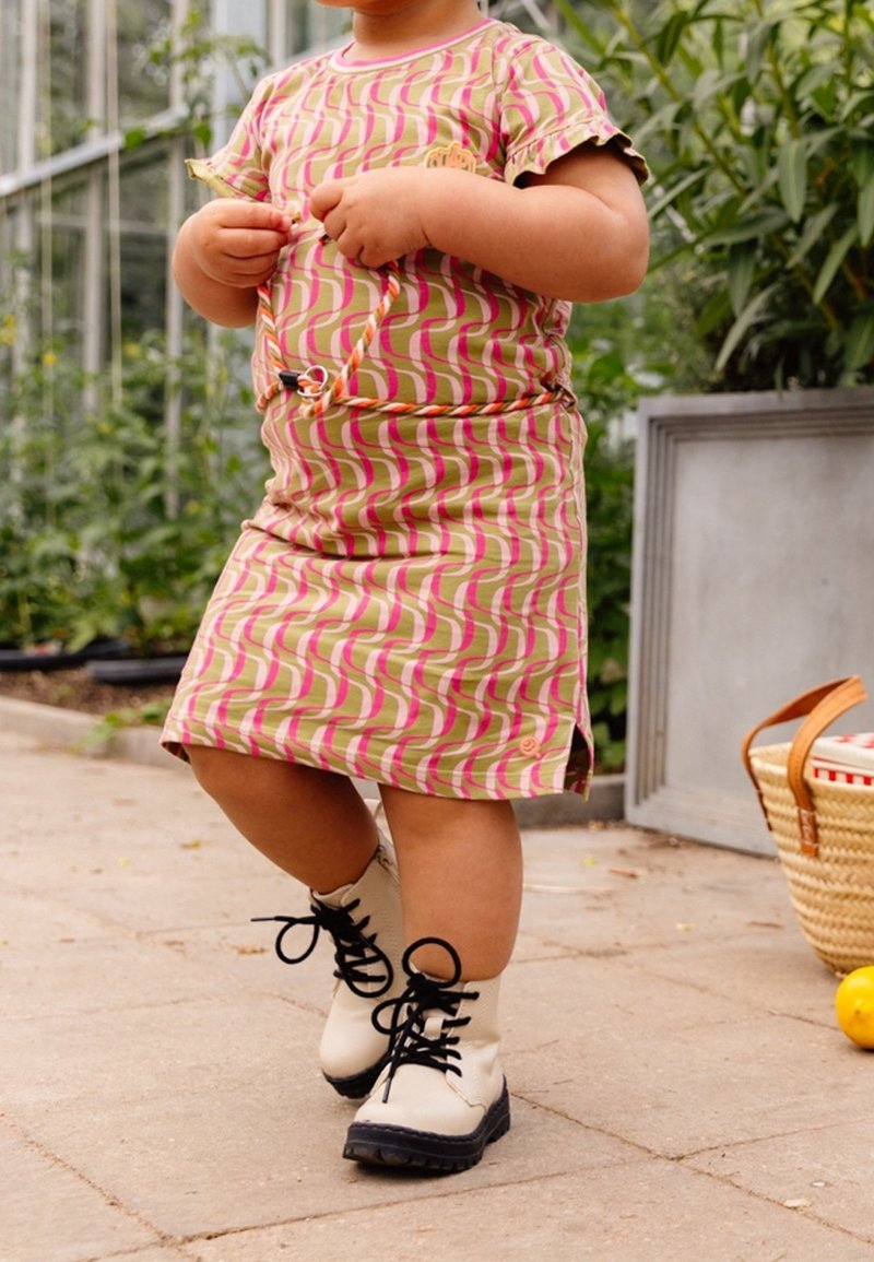 Niña con un vestido estampado y botas está de pie en un patio, sosteniendo una hoja con vegetación y una cesta en el fondo.