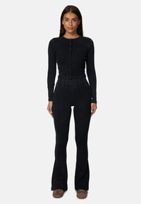 Zwarte lange mouwen cardigan met knopen, gecombineerd met nauwsluitende zwarte flare-pantalons. Beide gemaakt van een zachte, gestructureerde stof. Bruine schoenen.