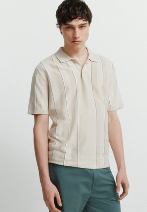 Junger Mann mit lockigem dunklem Haar, der ein beiges kurzärmeliges Strick-Poloshirt und türkisfarbene Hosen trägt, steht vor einem schlichten weißen Hintergrund.