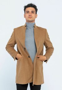 Brauner Wollmix-Mantel mit strukturiertem Design, einreihigem Verschluss und zwei vorderen Taschen, getragen über einem hellgrauen Rollkragenpullover.