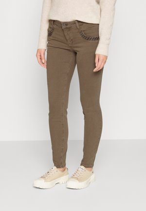 Frau trägt braune, schmal geschnittene Hose mit Vordertaschen und beige Sneakers mit dicker Sohle, stehend vor einfarbig hellem Hintergrund.