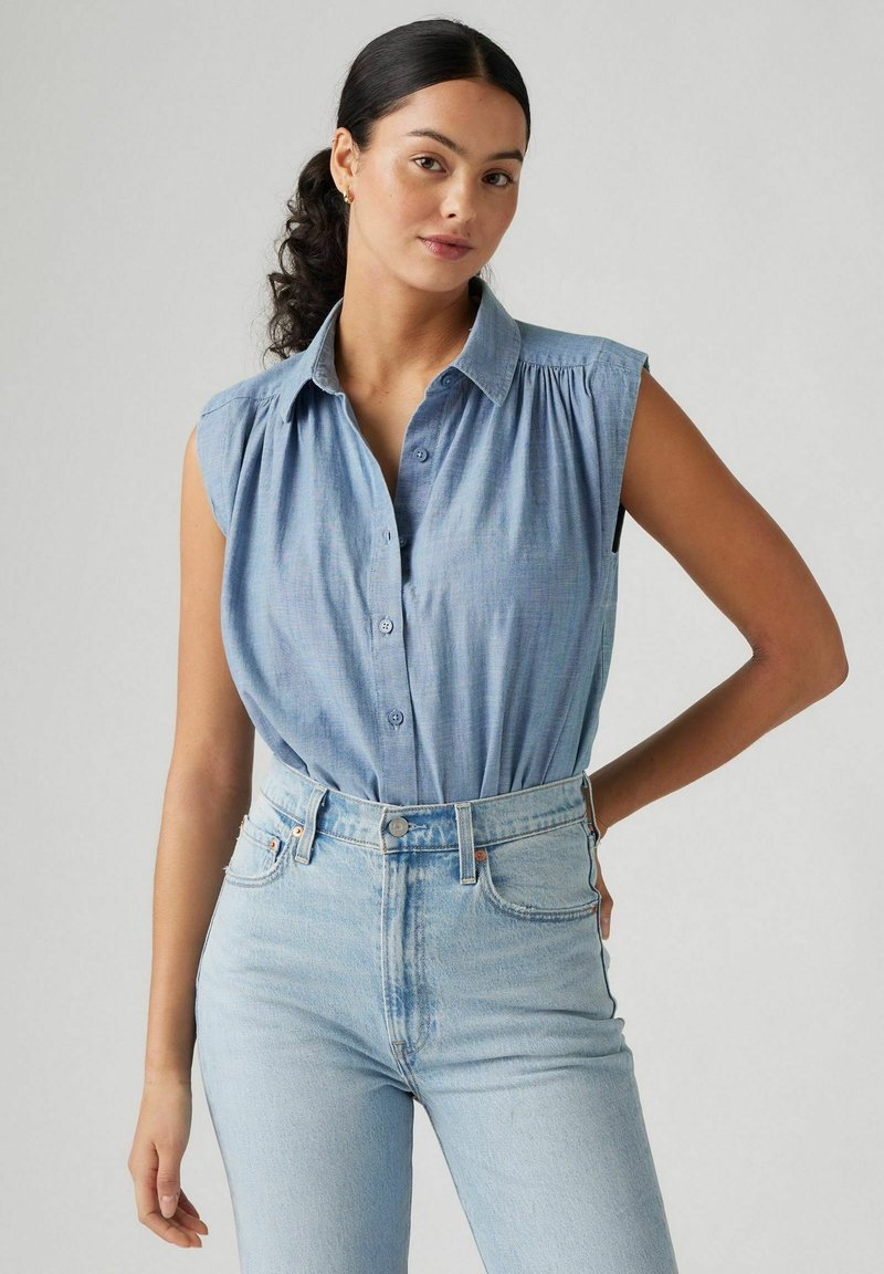 Chemise sans manches bleu clair avec un devant boutonné, en tissu doux et détails froncés aux épaules, portée avec un jean bleu clair taille haute.