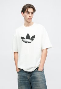 Biela voľná tričko s čiernym logom Adidas trefoil, so krátkymi rukávmi a okrúhlym výstrihom, v kombinácii s modrými džínami.