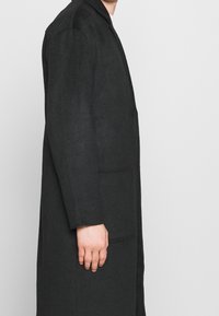 Persona che indossa un lungo cappotto nero con tasca applicata sul lato, in piedi di profilo contro uno sfondo chiaro e uniforme.