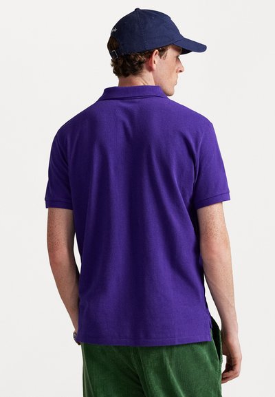 Camisa polo roxa com gola plana e mangas curtas; vestida com calças de veludo cotelê verde; vista traseira mostrando a textura do tecido e costuras.