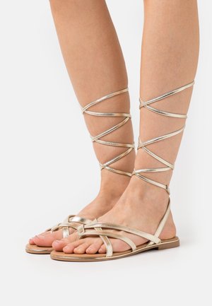 Fötter som bär platta, metalliska guld gladiator-sandaler med tunna remmar som sveper runt anklarna och underbenen mot en vit bakgrund.