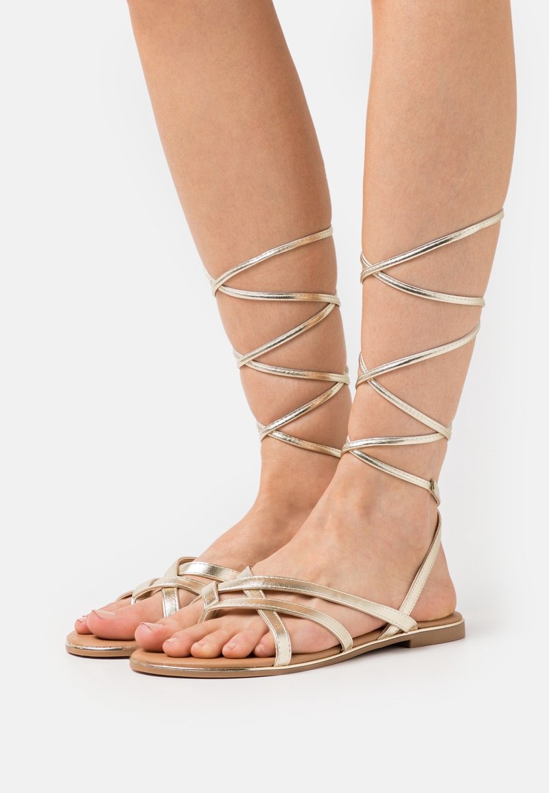 Jalat yang mengenakan sandal gladiator datar berwarna emas metalik dengan tali tipis mengelilingi mata kaki dan betis bagian bawah di latar belakang putih.