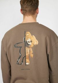 Sudadera marrón con un diseño gráfico de un oso de peluche y contornos, con el texto "AQUÍ ESTÁ JOHNNY" en la parte inferior. El tamaño y el ajuste son relajados.