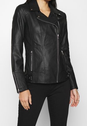 Femme portant une veste de motard ajustée en similicuir noir avec des panneaux matelassés, des poches zippées et des poignets zippés, associée à un pantalon noir.