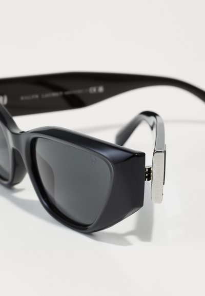 Schwarze Sonnenbrille mit einem geometrischen Rahmen, grauen Gläsern und silbernen Metallelementen. Die Bügel verfügen über eingravierte Markenbezeichnung und Scharnierelemente.