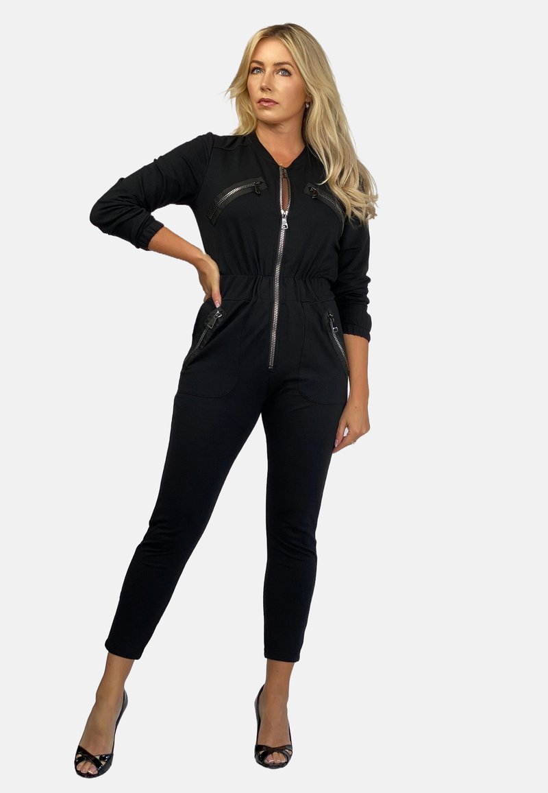 Czarny kombinezon wykonany z elastycznego materiału, z suwakiem z przodu, dwiema kieszeniami na zamek błyskawiczny na piersiach oraz zwężanymi nogawkami. Dodatki obejmują dopasowane mankiety i pas.
