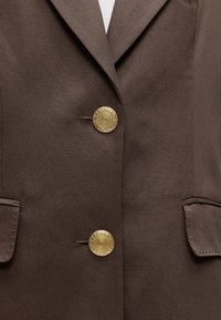 Καφέ σακάκι με δύο περίτεχνα χρυσά κουμπιά και δύο τσέπες με καπάκι, εμφανίζεται σε έναν κορμό με λευκό εσωτερικό πουκάμισο.