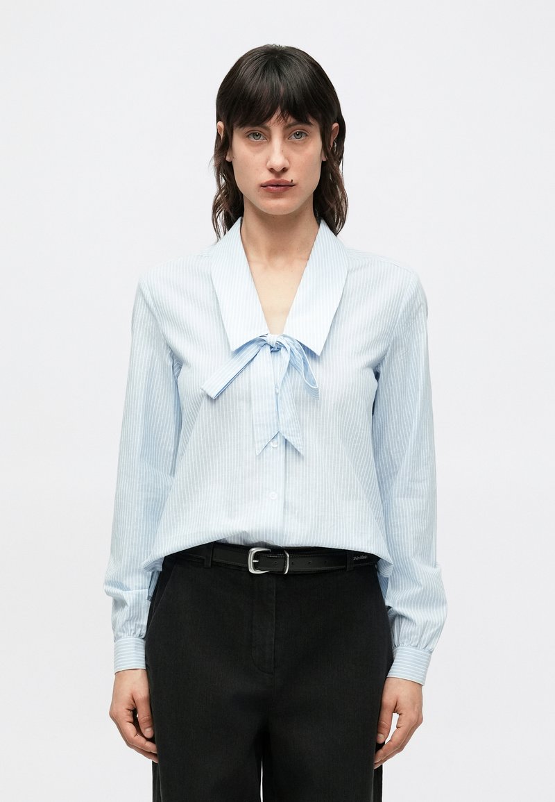 Svetlomodrá pásikavá blúzka s veľkým golierom a detailom viazanky; dlhé rukávy; v kombinácii s tmavými nohavicami a čiernym opaskom.