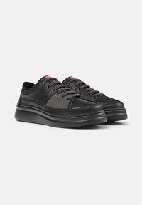 Zwarte vetersneakers met verschillende texturen en een dikke zool, voorzien van een klein rood logo nabij de tong. Gepresenteerd tegen een neutrale achtergrond.