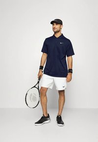 Man draagt een marineblauw polo, witte korte broek, zwarte zonneklep, polsbandjes en sneakers, en houdt een tennisracket vast tegen een effen achtergrond.
