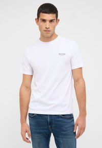 Biały bawełniany T-shirt z okrągłym dekoltem i krótkimi rękawami. Na lewej piersi znajduje się małe szare nadrukowane logo. Noszony z niebieskimi jeansami.