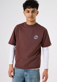Camiseta marrón de manga corta con cuello redondo y detalle de logo circular azul en el pecho, llevada sobre una camisa blanca de manga larga.