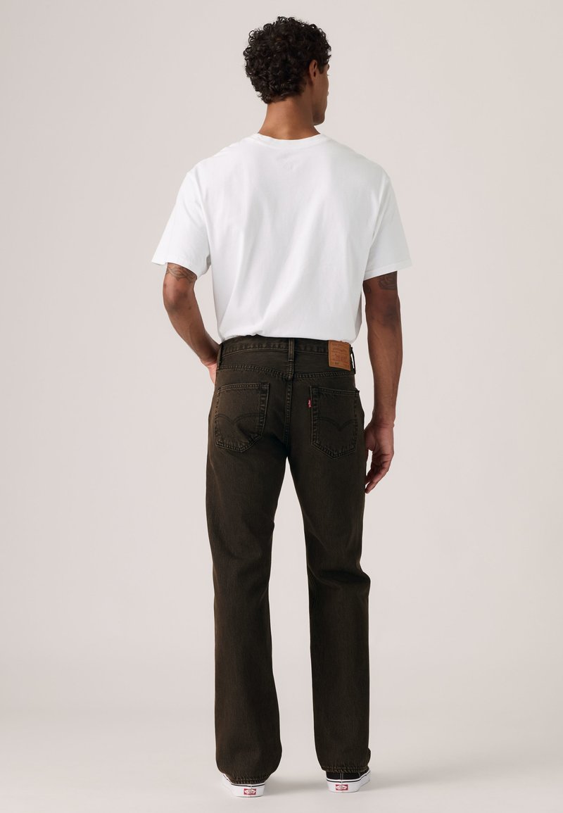 Mand med krøllet hår iført en ensfarvet hvid t-shirt og mørkebrune jeans, står med ryggen til og har den ene hånd på hoften.