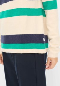 Beige Pullover mit marineblauen und grünen Streifen, strukturiertem Strick, lässiger Passform, kombiniert mit dunklen Hosen. Etikett an der Seitennaht sichtbar.